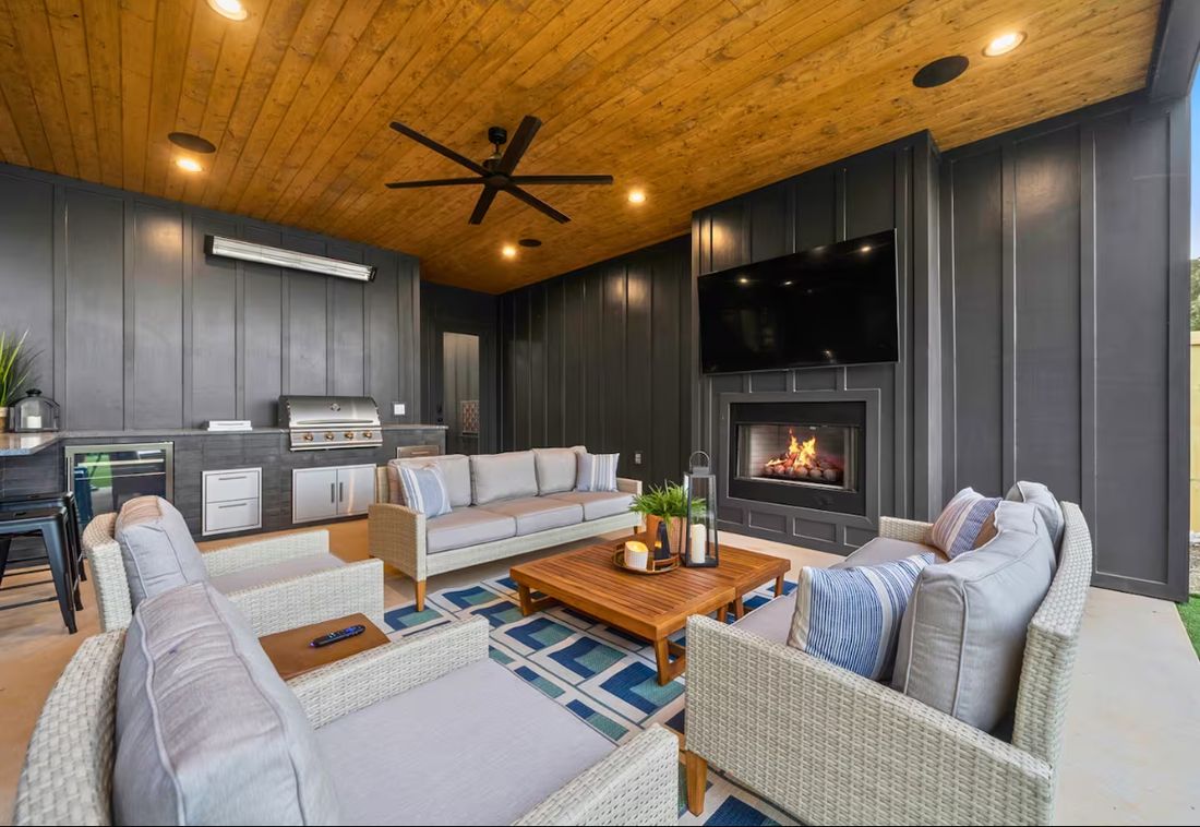 Covered outdoor living area with a built-in grill and outdoor kitchen, gas fireplace, dark board-and-batten walls and a stained wood plank ceiling.
