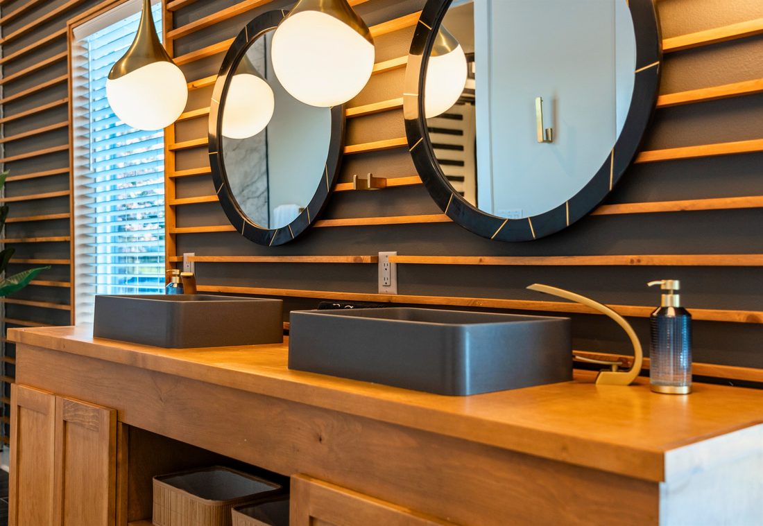 Bathroom vanity with a wood slat feature wall, a live-edge wood countertop, two charcoal vessel sinks, round mirrors and brass pendant lights.