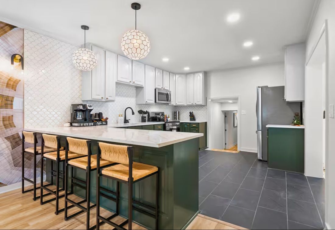 Kitchen peninsula with green cabinetry, quartz waterfall countertop, woven bar stools, scalloped tile backsplash and globe pendant lights.