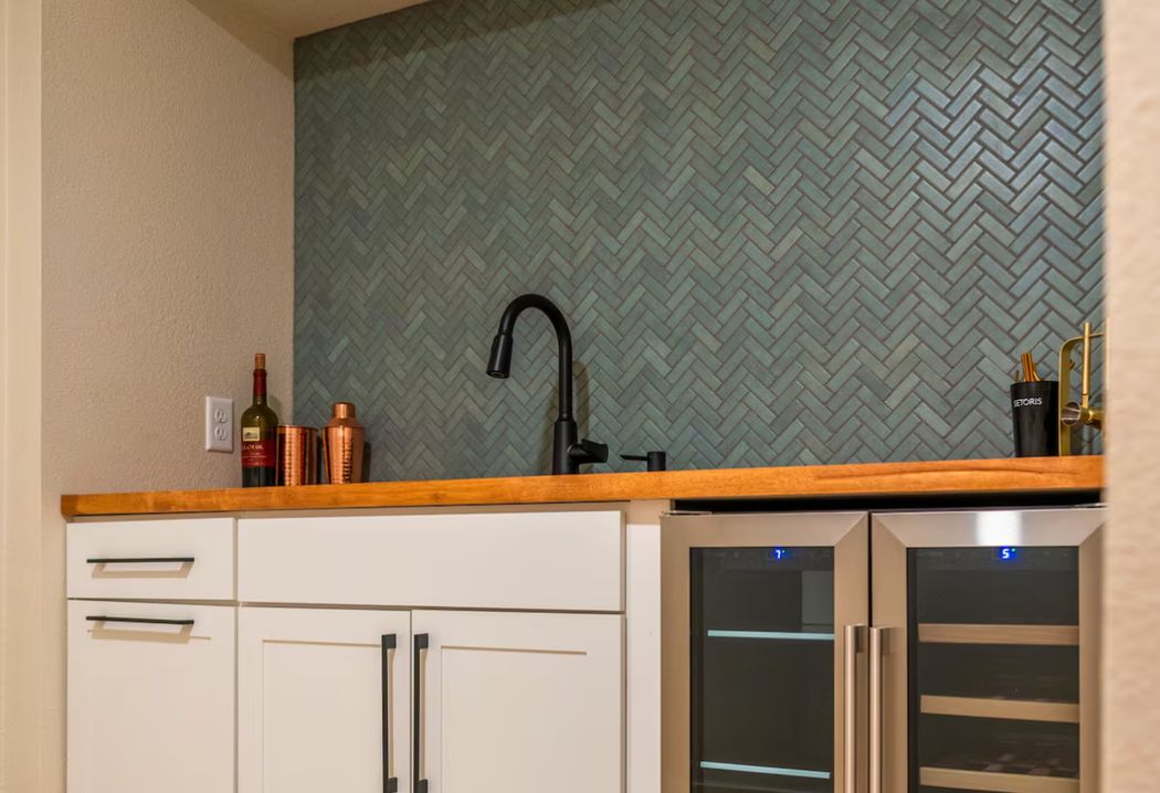 Wet bar with white shaker cabinets, a live-edge wood countertop, teal herringbone tile backsplash, matte black faucet and two wine refrigerators.