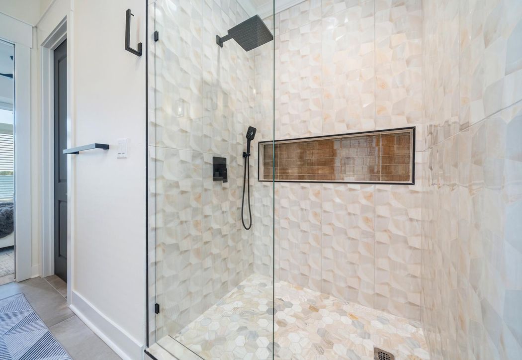 Walk-in shower clad in textured three-dimensional cream stone tile with a wood-look tiled niche, matte black fixtures and a marble hexagon floor.