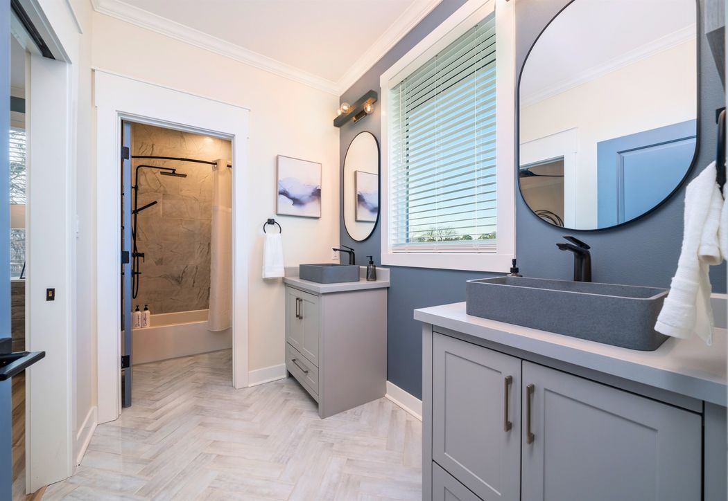 Bathroom with two grey vanities, stone vessel sinks, round black mirrors, a blue accent wall and a herringbone tile floor.